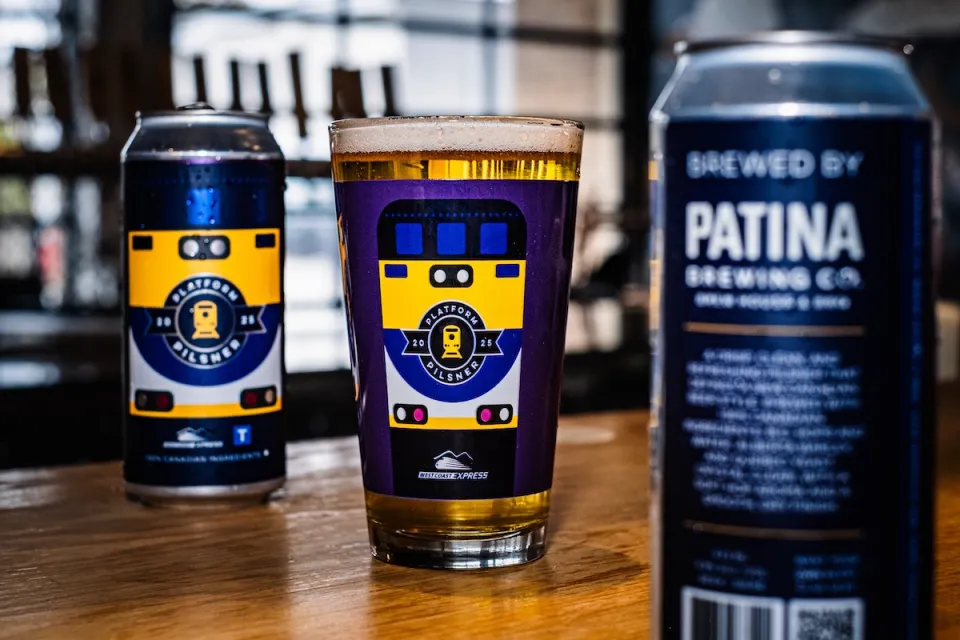 Close-up of full beer glass with purple train-themed design sitting on bar between two frosty tall cans.